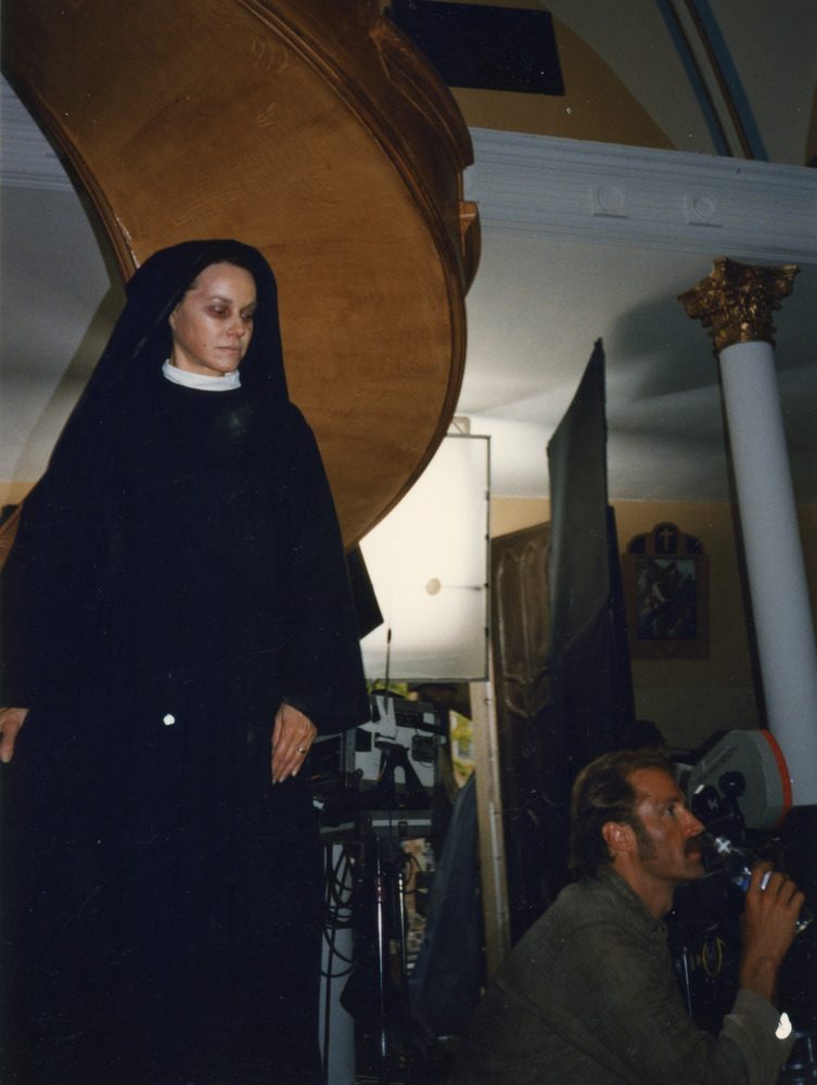 Woman in a habit stands on the circular staircase, with the filming set around her.