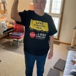 An older white woman wearing a yellow and black "Save Bernheim Now" t-shirt in a rosie the riveter pose with her arm.