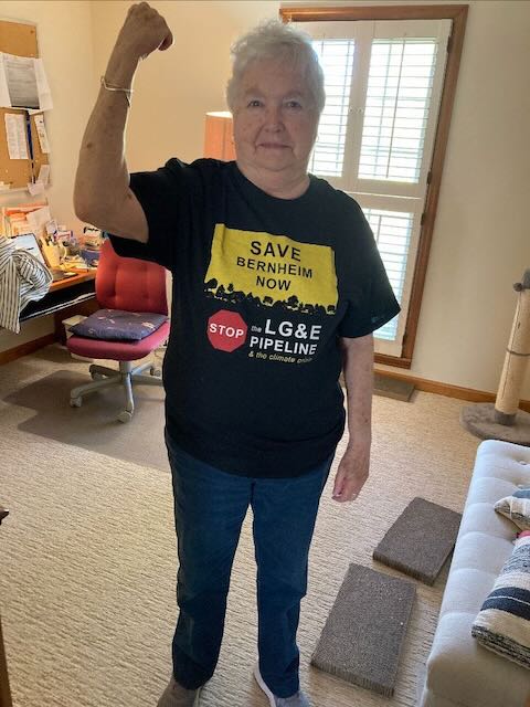 An older white woman wearing a yellow and black "Save Bernheim Now" t-shirt in a rosie the riveter pose with her arm.