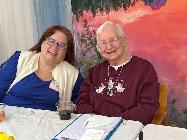 Two women smiling for a photo sitting at a table, the women on the left has a blue shirt and longer brown hair, the woman on the right has a burgendy sweater with angels on it and short white hair.