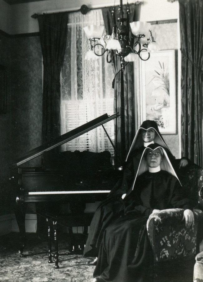 Archival photo of two sisters in habits sitting in a parlor next to the piano.
