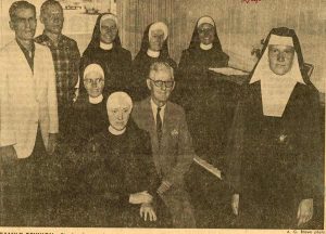 Archived newspaper clipping showing a family posing for a photo around the piano. There are five women in one style of habit, one wearing a different type of habit, two men standing to the left of the photo and one older man sitting on the piano bench.