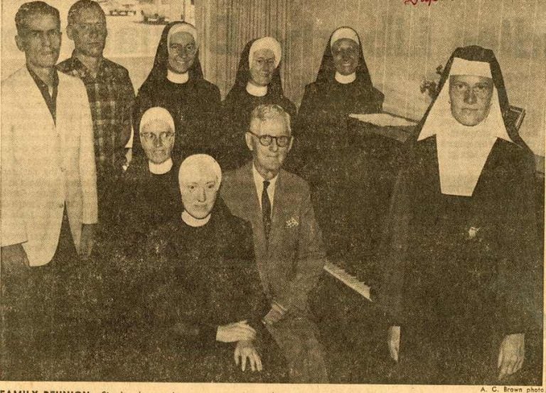 Archived newspaper clipping showing a family posing for a photo around the piano. There are five women in one style of habit, one wearing a different type of habit, two men standing to the left of the photo and one older man sitting on the piano bench.
