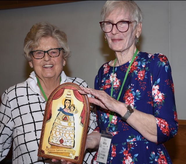 From left, an older white woman with black glasses and a striped black and white t-shirt stands next to an older white woman with brown glasses and a floral blue and pink dress. They are holding a plaque with biblical art of mother Mary and baby Jesus.