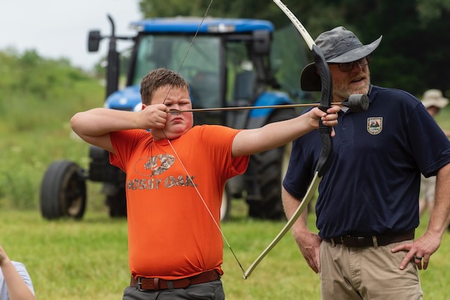 A kid in an orange shirt focusing intently on shooting a bow and arrow.