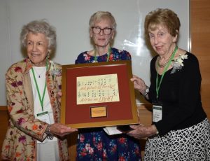 Three older white women in nice colorful clothing pose for a picture with a plaque that has music notes on it.