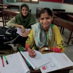 Photo of two Pakistani middle aged girls wearing colorful clothing, one in green patterns and the other with yellow, red, green and black patterns smiling as they take a break from working on their school work which is displayed on the desk in front of them.