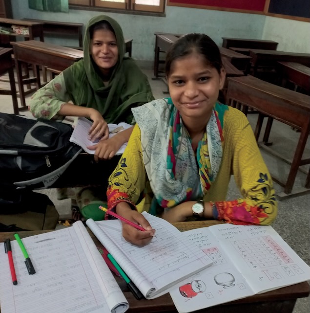 Photo of two Pakistani middle aged girls wearing colorful clothing, one in green patterns and the other with yellow, red, green and black patterns smiling as they take a break from working on their school work which is displayed on the desk in front of them.