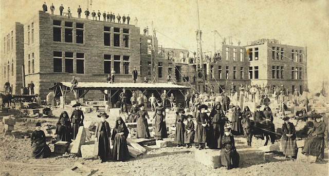 Sepia toned older photograph of many people in habits and black clothing standing in front of the Loretto Heights Academy construction site, a building is half finished and hollow in the background.