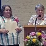 Two women standing together, the woman to the right is standing at a podium speaking into a microphone. She has short, grey hair, and glasses, and a bouquet of pink and yellow roses, and babies breath sits directly in front of the podium. The woman to the left has longer, brown hair, is wearing a blue and white striped blouse, with a pink boutonniere, holding a beige filing folder in her hands.