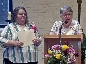 Two women standing together, the woman to the right is standing at a podium speaking into a microphone. She has short, grey hair, and glasses, and a bouquet of pink and yellow roses, and babies breath sits directly in front of the podium. The woman to the left has longer, brown hair, is wearing a blue and white striped blouse, with a pink boutonniere, holding a beige filing folder in her hands.