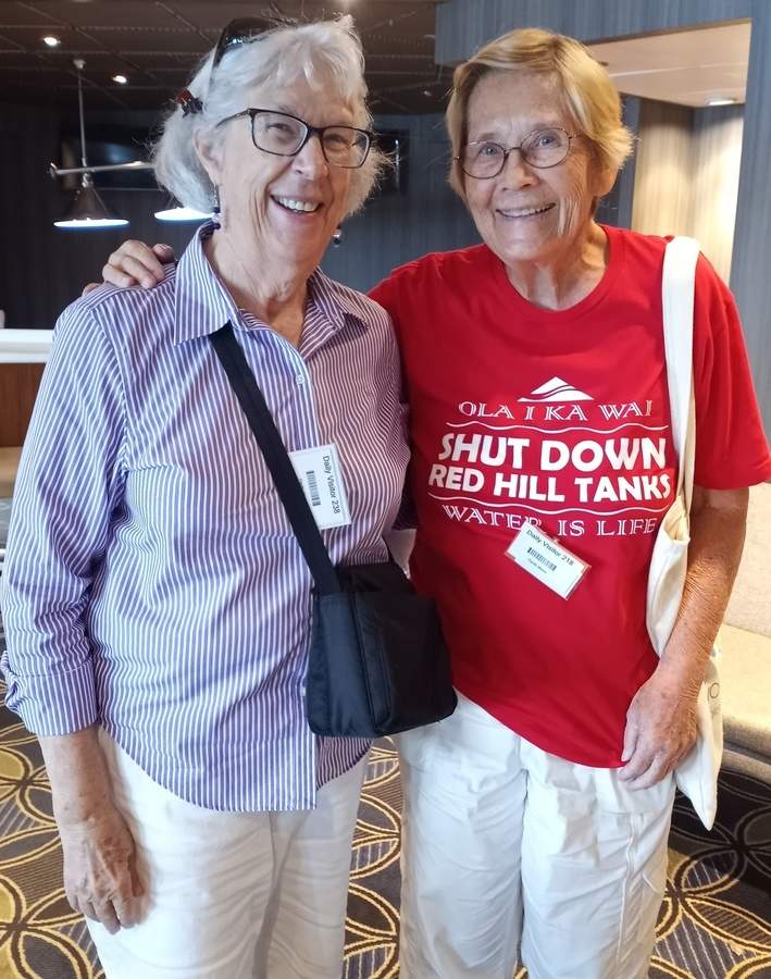 Two women pose together in a room for a photo. The woman on the right wears a red shirt that reads "Olaika Wai. Shut Down Red Hill Tanks. Water is Life."