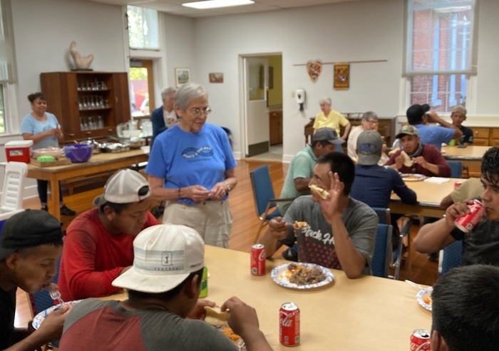 A woman with short, grey hair and glasses, wearing a blue shirt standing up conversing with a table of men eating a meal in an indoor cafeteria.