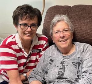 A woman with short, grey hair and glasses, wearing a grey blouse and sitting in a brown recliner chair, accompanied by a woman with short, brown hair, black rimmed glasses, wearing a red and white striped, collared shirt and a black name tag. Both women are smiling for a picture together indoors.