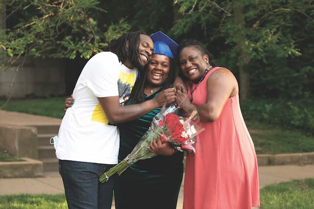 A Marian Middle School graduate is pictured in the middle of an embrace from a family member on each side of her as she holds a bouquet of flowers.