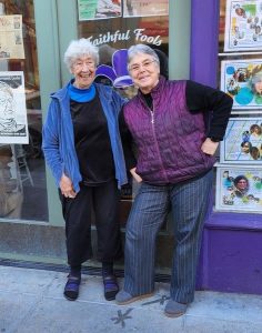 Two friends smile together for a picture in front of a purple building with green doors outside. Both friends are women with short, grey hair, one is laughing, and the other with glasses is posing silly with her hand on her hip.