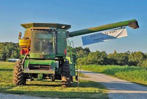A large green tractor in a bright green field on a clear blue-sky day. The tractor has an arm extended to its left, where a banner stating, "Welcome to the Ag Bash" is displayed over a gravel pathway.