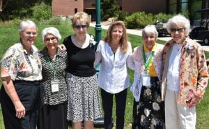 Six women standing and smiling together for a group picture outdoors on a sunny day in front o a green grass field and a dark green lamp post. They are all wearing nice dress clothing, and two women are wearing matching green lanyards with a white card on the end.