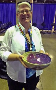 A woman with short grey hair and glasses, wearing a blue blouse, a white unbuttoned collared button up shirt, and a colorful lanyard smiling brightly with her eyes closed, holding a bowl, in an event space with chairs and blue curtains indoors.