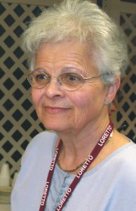 A woman with metal framed glasses and white hair wearing stud silver earrings giving a soft smile not looking at the camera. She is wearing a blue shirt with a lanyard that has the word "Loretto" repeating.