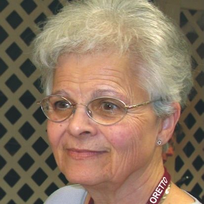 A woman with metal framed glasses and white hair wearing stud silver earrings giving a soft smile not looking at the camera. She is wearing a blue shirt with a lanyard that has the word "Loretto" repeating.