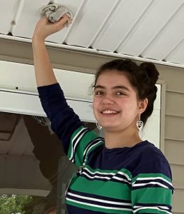 A girl in a horizontal striped green, blue and white shirt smiles for a picture as she cleans a ceiling panel with a white rag.
