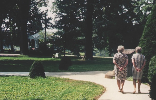 Two women in patterned dresses walking together on a paved path in between green grass, trees and shrubs.