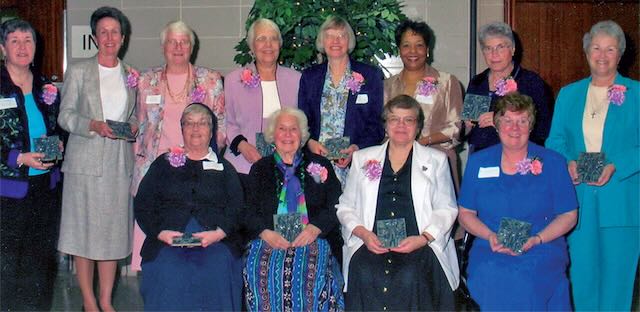 A group of 12 women half sitting half standing wearing nice colorful clothing, pink and purple corsages and holding an award.