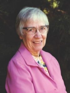 An older woman with glasses and white hair photographed smiling wearing a pink blazer. She is standing in front of lush pine tree branches.