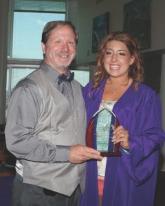 A man in a grey suit takes a smiley photo with a high school student in a purple graduation gown while they hold up a translucent award together.