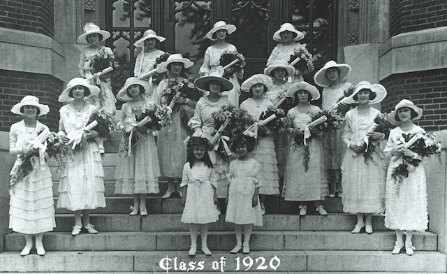 A Loretto College class picture circa 1920 where 15 adult women and two little girls stand staggered on school steps wearing white dresses, white hats, and each holding a bouquet of flowers.