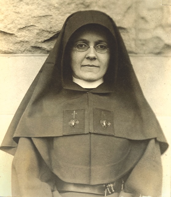 An older sepia toned photo of a nun in a habit with small round glasses looking at the camera.