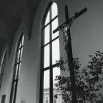Black and white photo of a crucifix in the foreground lit by light from tall church windows behind it.