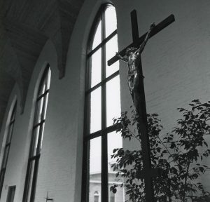 Black and white photo of a crucifix in the foreground lit by light from tall church windows behind it.