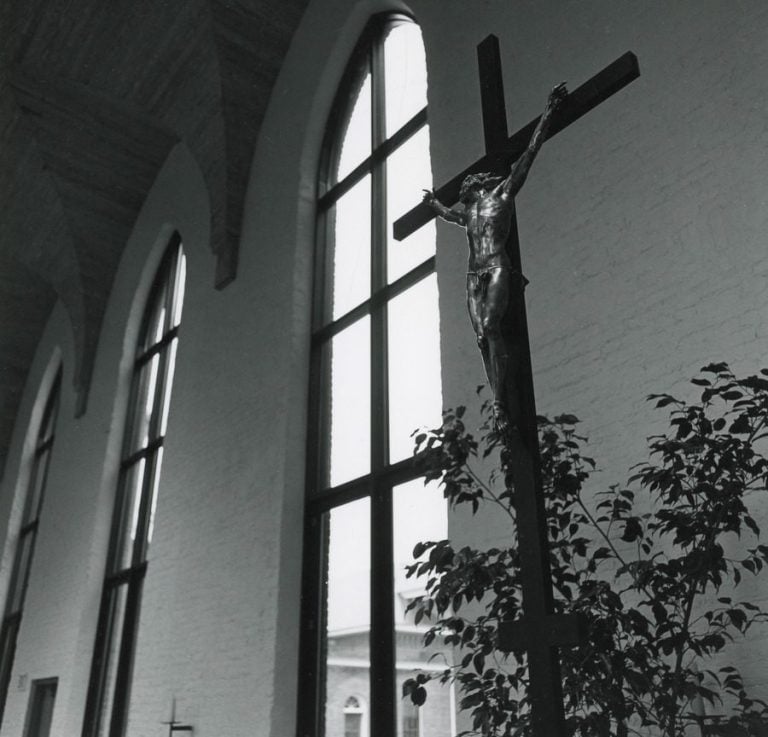 Black and white photo of a crucifix in the foreground lit by light from tall church windows behind it.
