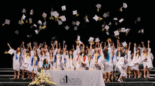 A large group of Loretto Academy seniors in their white graduation caps and gowns tossing their caps at the commencement ceremony.