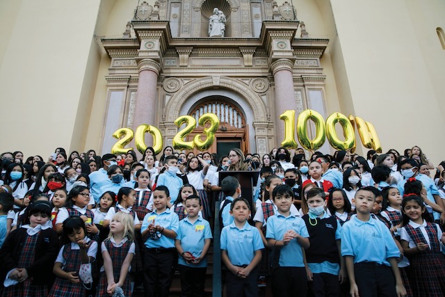 A group of young students in their school uniforms stand outside on the Loretto Academy school steps to celebrate their school's 100-year anniversary. There are gold balloons above the crowd "2023" and 100th"