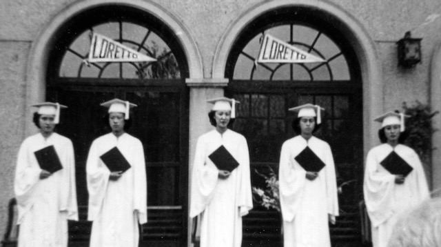 A black and white photo of five women in white graduation cap and gowns holding their diplomas in front of two doors that have "Loretto" signs on them.