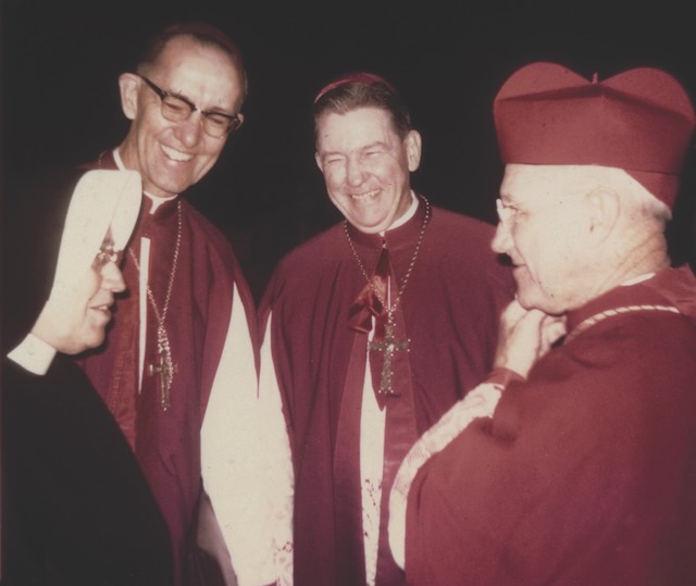 A nun in a black habit talking with two Catholic bishops and one cardinal all dressed in red religious gowns.