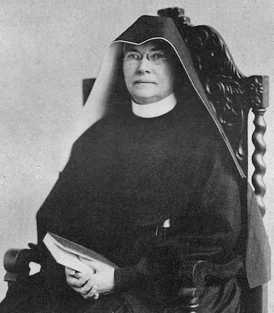 A black and white photo of a nun in a full habit and glasses holding a book sitting in an ornate chair.