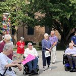An outdoor ceremony event on a sunny day with large green trees and a big brick building in the background. Many attendees are standing, some are in wheel chairs, and two women in wheelchairs have neon pink papers and a microphone.