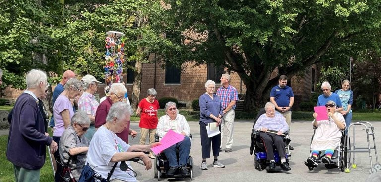 An outdoor ceremony event on a sunny day with large green trees and a big brick building in the background. Many attendees are standing, some are in wheel chairs, and two women in wheelchairs have neon pink papers and a microphone.