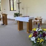A newly renovated altar space with light grey carpet, white brick walls, and wooden furniture and a large cross.