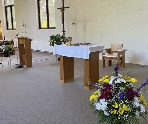 A newly renovated altar space with light grey carpet, white brick walls, and wooden furniture and a large cross.