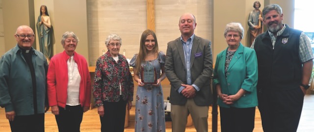 Seven people in a row taking a photo with an award recipient, a young woman with long light brown hair in a blue patterned dress in the center.