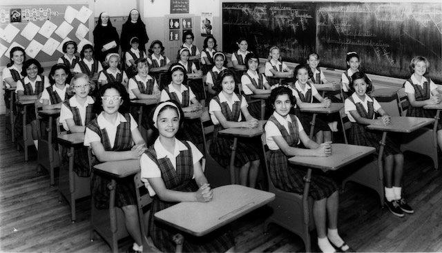 Students sitting attentively at their desks in 4 rows with two nuns in full habits standing in the back of the classroom.