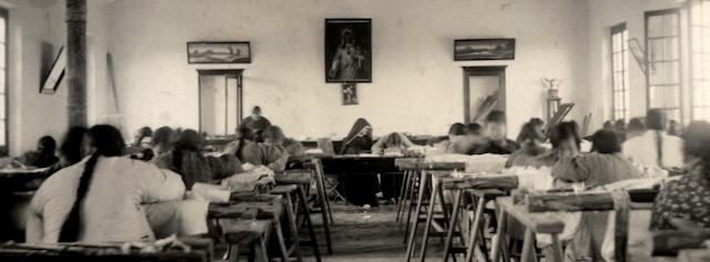 A sepia toned photo taken from the back of a classroom showing two rows of students at desks practicing embroidery and a nun instructing from her desk at the front of the room in between the two rows.