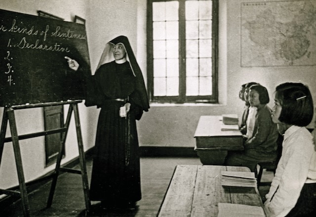 Black and white photo of a nun at a chalkboard teaching to an attentive class of students.