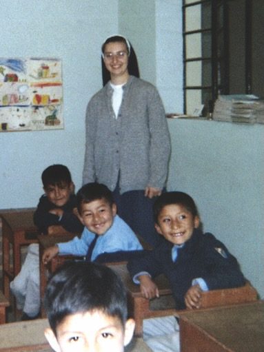 A young nun wearing glasses, habit and grey sweater stands behind four kids sitting at desks at a school in Tacna, Peru.