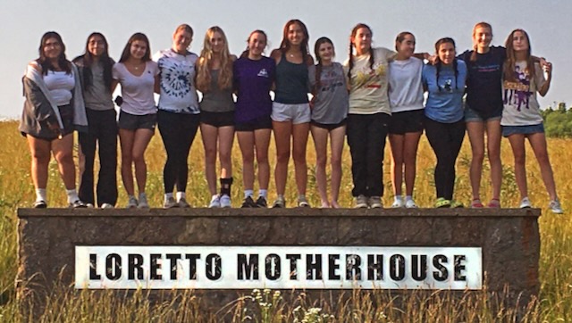 A group of 13 girls stand in a row on top of a Loretto Motherhouse sign in the middle of tall grass.